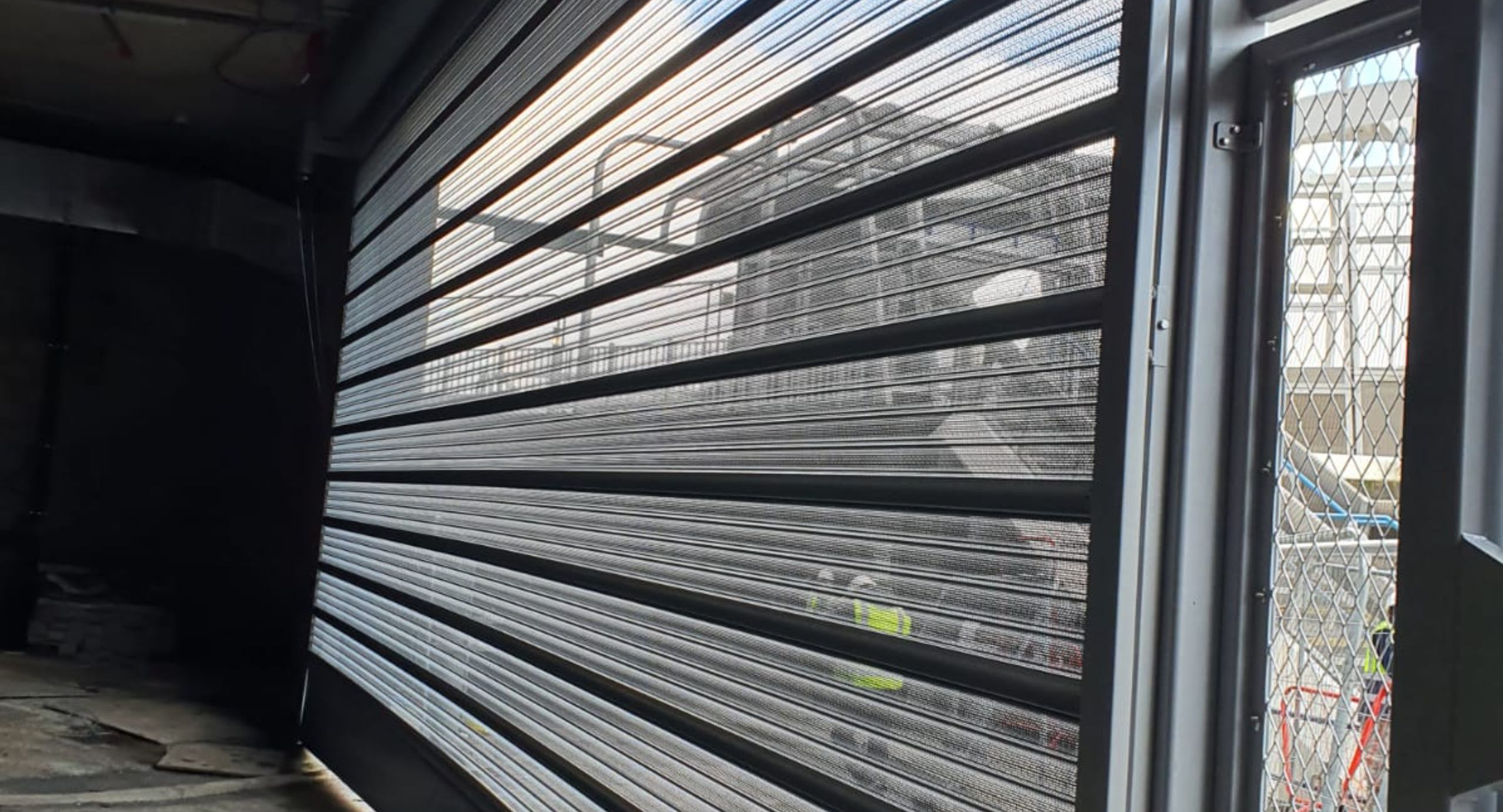 A large, dark grey perforated roller security shutter is shown partially open, angled from the bottom left to the right side of the frame. The shutter features horizontal slats with numerous small perforations, allowing diffused sunlight and a view of an outdoor construction site with chain-link fencing to pass through. The interior space, visible to the left and bottom, is a raw concrete area, suggesting an industrial or commercial setting like a garage or loading dock.