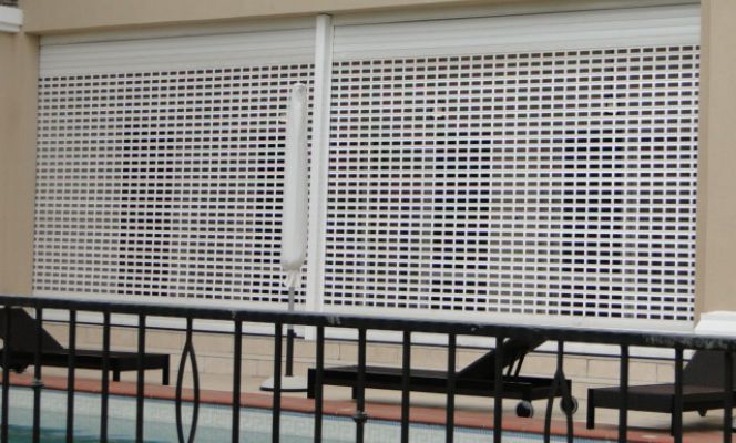 A building facade showcases two wide white perforated roller security shutters with a pattern of small rectangular openings. The shutters cover the ground-level opening, with a light beige wall and a balcony with a black railing visible above. In the foreground, a dark metal railing and a swimming pool area with lounge chairs are partially obscuring the lower part of the shutters.